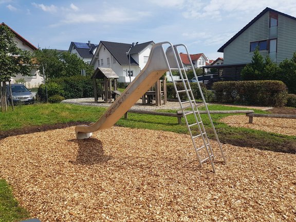 Das Foto zeigt den Blick auf einen Spielplatz in einem Wohngebiet. Zentral ist eine Rutsche zu sehen, im Hintergrund erkennt man einen Spielturm aus Holz. Noch weiter im Bildhintergrund sieht man unter blauem Himmerl mehrere Wohnhäuser.