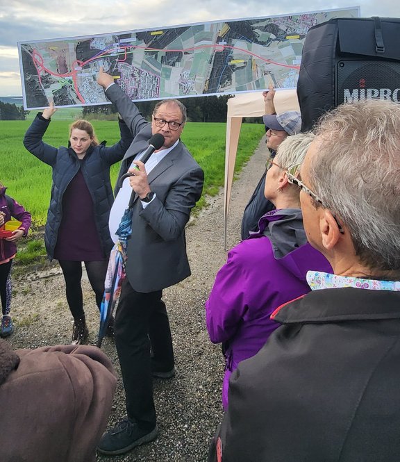 Eine Gruppe von Personen. Einige sind von hinten zu sehen. In der Mitte steht der Oberbürgermeister und zeigt mit seinem Schirm auf einen Plan. Die Gruppe ist auf einem Feldweg und schaut sich vor Ort um. Grund ist der Ausbau der Straße.