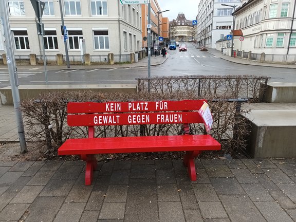 Die Bank ist rot gestrichen und darauf ist weißer Farbe geschrieben: Kein Platz für Gewalt gegen Frauen. Sie steht auf einem Platz an einer großen Kreuzung in Schwenningen.