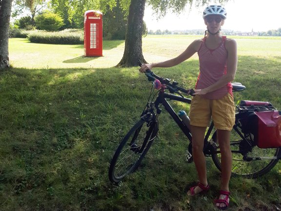 Rechts eine Fahrradfahrerin unter einem Baum im Schatten auf einer grünen Wiese