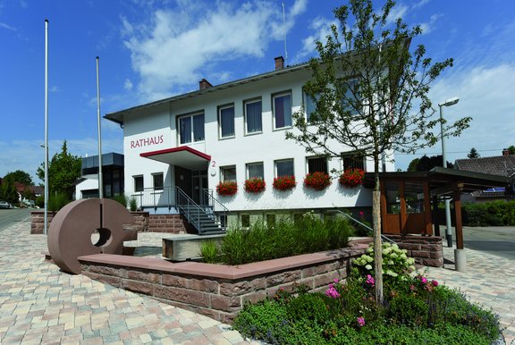 Rathaus Obereschach von vorne. Weiße Fassade mit Blumenkästen in dem rote Blumen sind. Im Vordergrund ist der neue Sandsteinbrunnen zu sehen, welcher rund ist. Rechts vom Bild ist noch die Bushaltestelle vor dem Rathaus zu sehen.