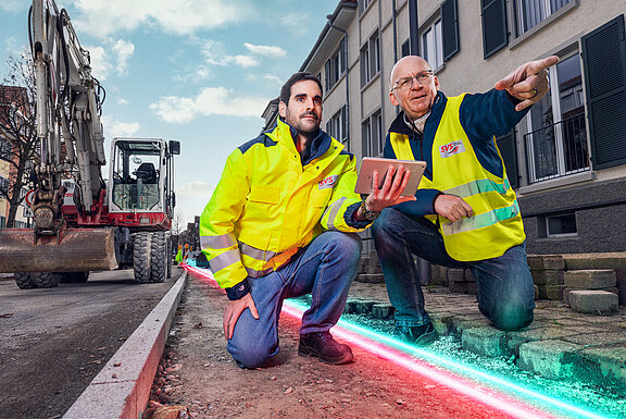 Zwei Männer in einer gelben Arbeitsweste. Beide knieen am Boden in einer Baustelle. Der rechte Mann zeigt mit der Hand nach vorne.