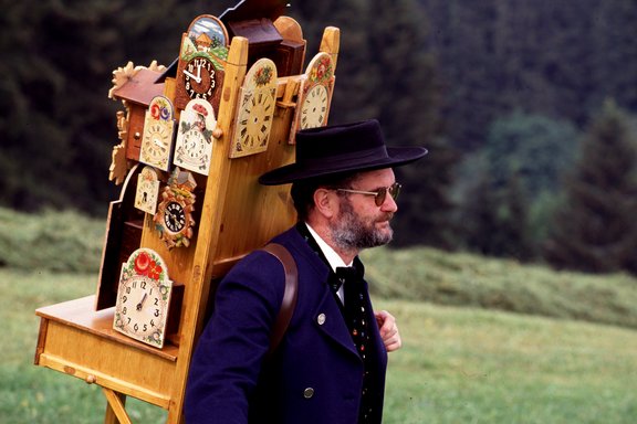 ein Mann, traditionell in Tracht gerkleidet, trägt ein Holzgestell mit Uhren auf dem Rücken