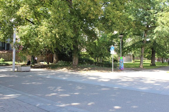 Im Vordergrund ist die Ringanlage, Ecke Paradiesgasse und Kaiserring, zu sehen. Dahinter stehen große Bäume. Seitlich der Bäume stehen zwei Straßenlaternen. Auf einer Bank sitzt ein Mann. Ein weiterer Mann steht etwas abseits an der Stadtmauer.