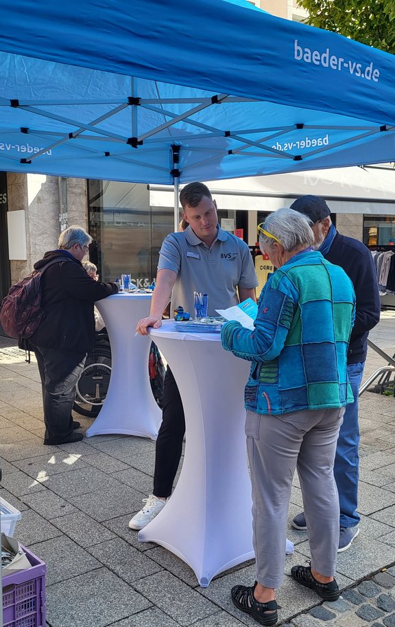 Unterschiedliche Personen sind an einem Stand in der Innenstadt und stehen an Tischen und unterhalten sich.