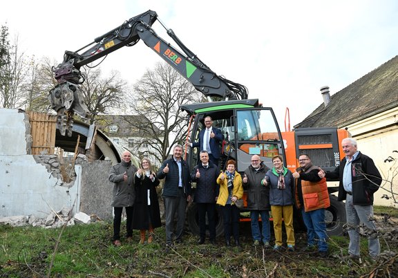 Männer und Frauen stehen vor einem Bagger und zeigen den Daumen hoch. Der Bagger hat eine alte Halle abgerissen. Dort werden neue Wohnungen gebaut.