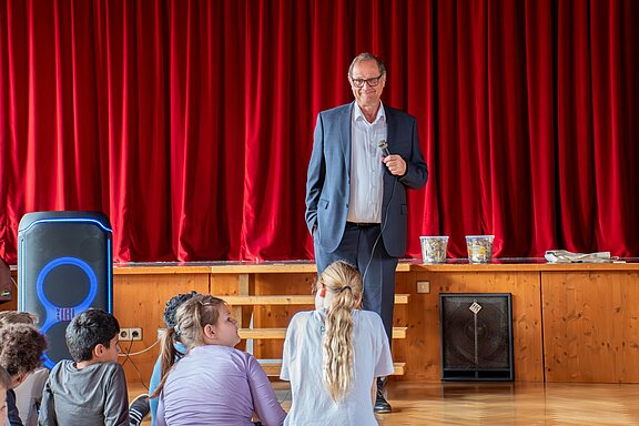 Der OB steht vor einem roten Vorhang in der Aula. Kinder sitzen vor ihm und stellen ihm viele Fragen.