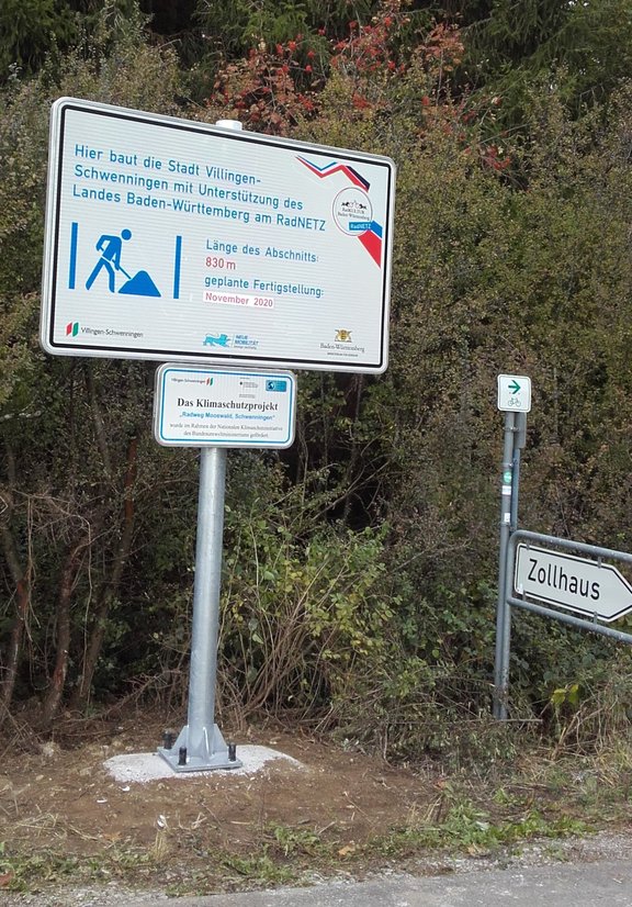 Großes weißes Schild mit kleinerem darunter. Im Hintergrund sind Büsche.