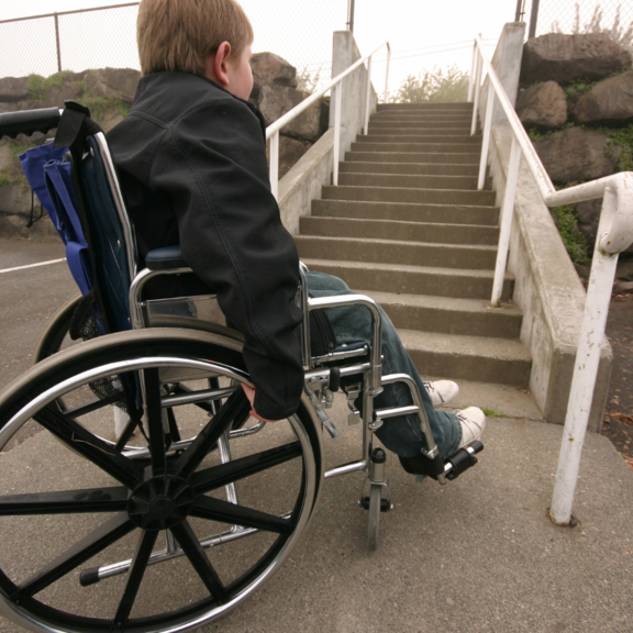 Kleiner Junge sitzt im Rollstuhl und steht vor einer Treppe. Es besteht nicht die Möglichkeit die Treppe mit dem Rollstuhl zu bewältigen.