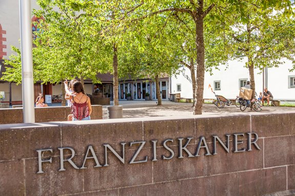 auf der Mauer vor dem Kulturzentrum, ist der Schriftzug Franziskaner zu lesen. Im Hintergrund sitzen Personen auf der Mauer und essen ein Eis.