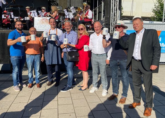 Acht Personen, sieben Männer und eine Frau, stehen nebeneinander vor einer Bühne, auf welcher eine Musikgruppe Musik spielt. Die acht Personen halten je ein Bierglas in die Höhe.